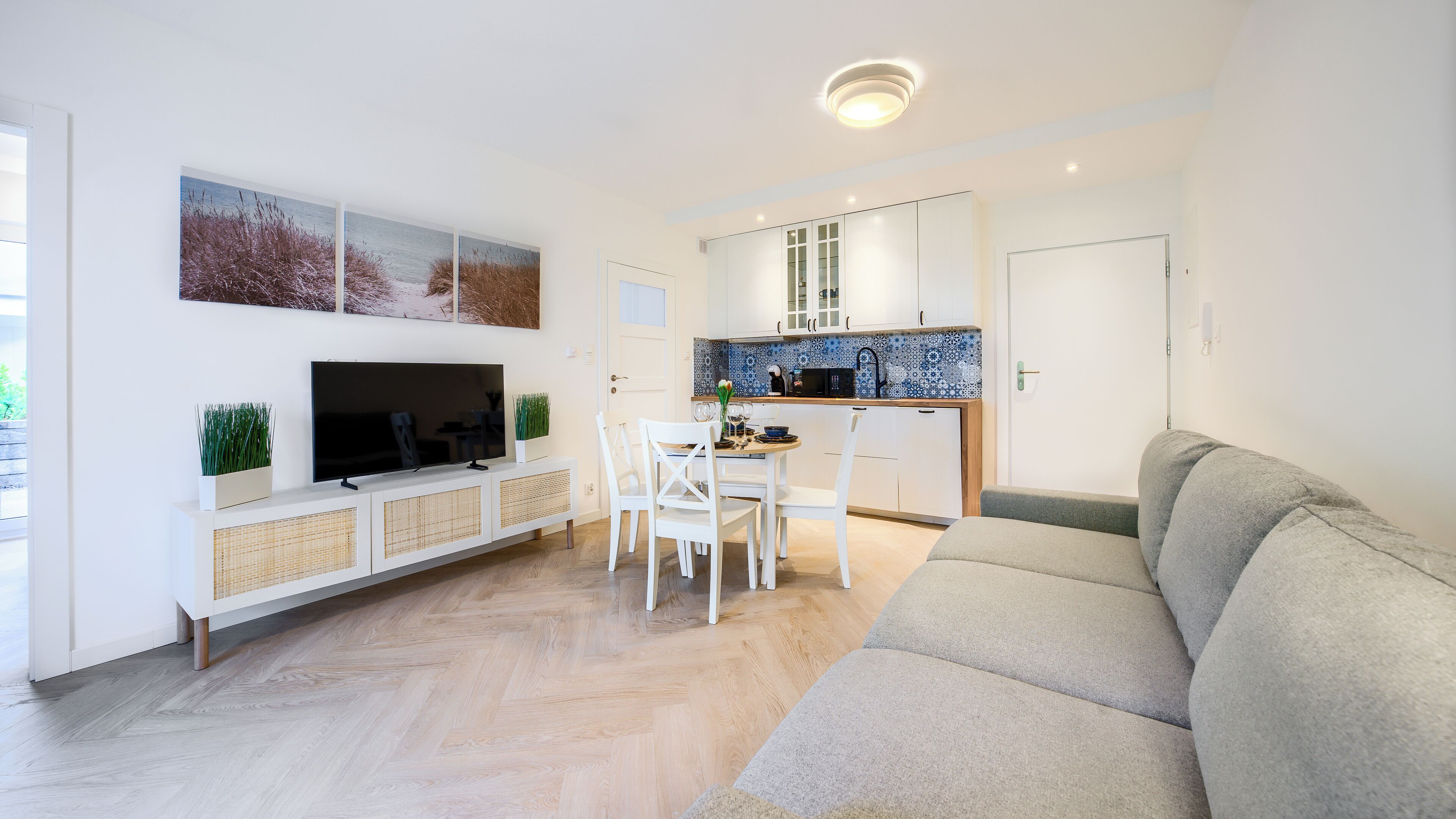 A modern living room with a flat-screen TV, white furniture, a dining area, and a kitchen with patterned backsplash.