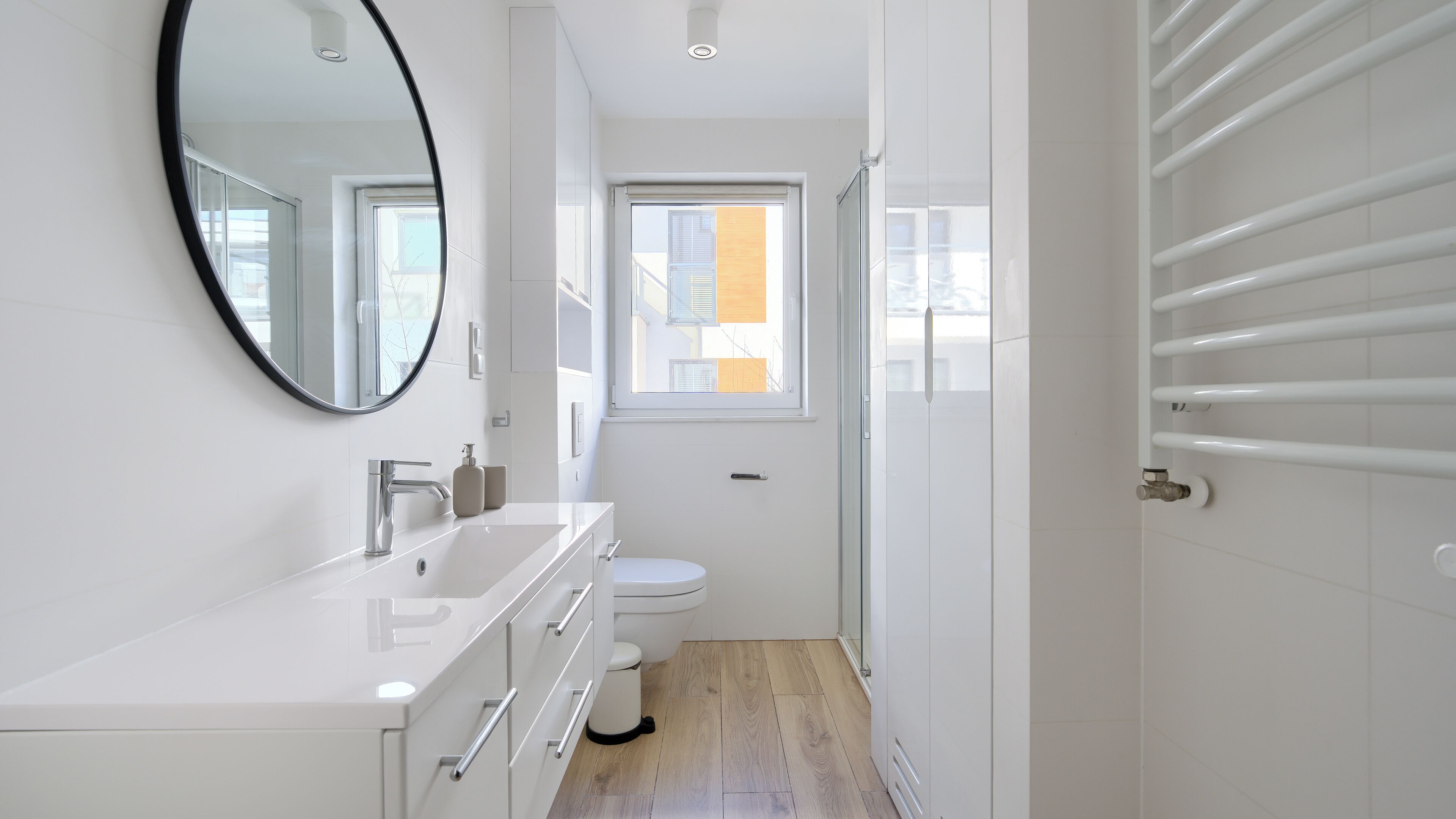 A modern bathroom with a white vanity, a round mirror, a toilet, and a window.