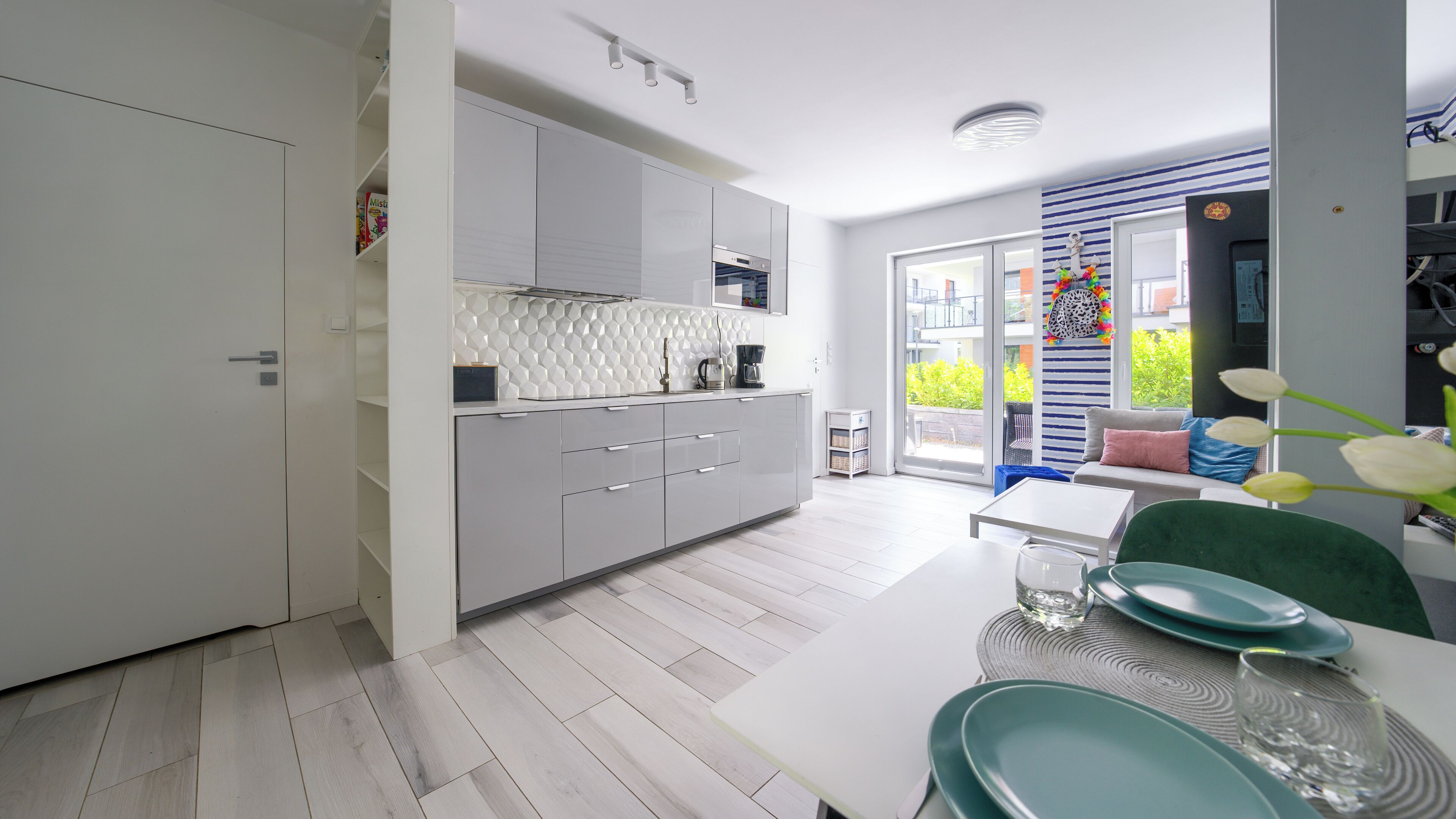 A modern kitchen with white cabinets, a dining area with a white table and teal chairs, and a view of greenery through the glass door.