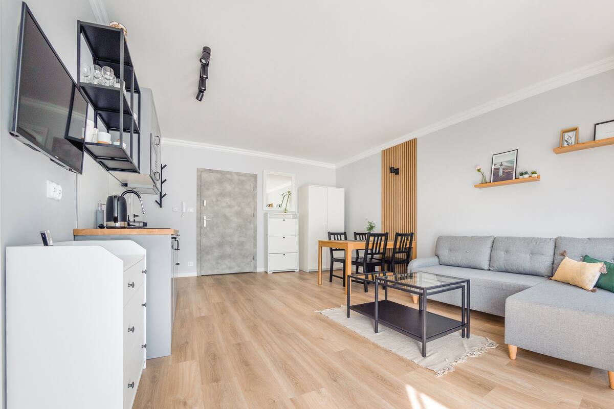 Un salon moderne avec un canapé gris, une table à manger avec des chaises, une table basse, un téléviseur fixé au mur et un coin cuisine équipé d’un réfrigérateur et d’un four à micro-ondes.