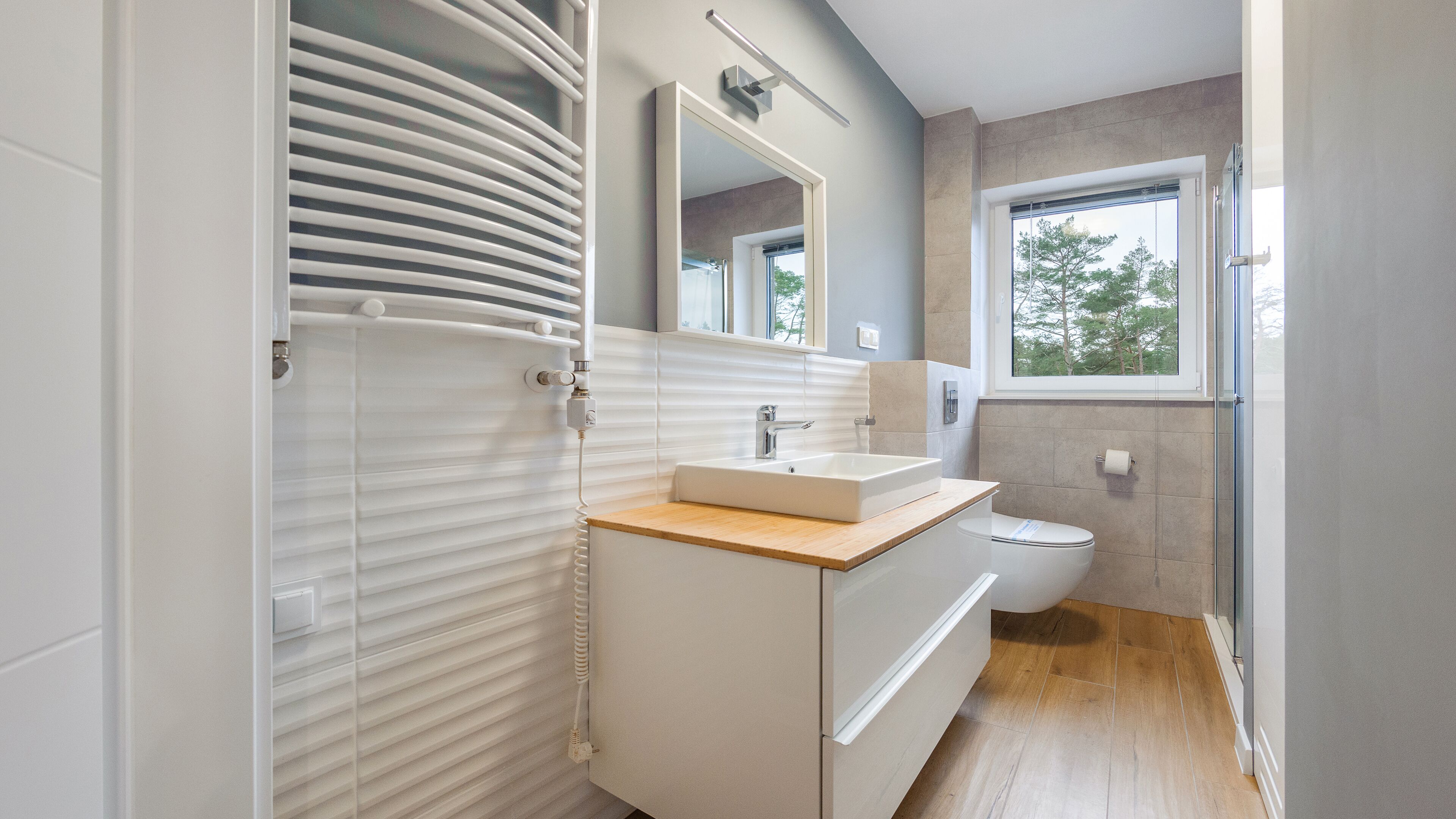 A modern bathroom with a white vanity, a mirror, a toilet, and a window with a view of trees.