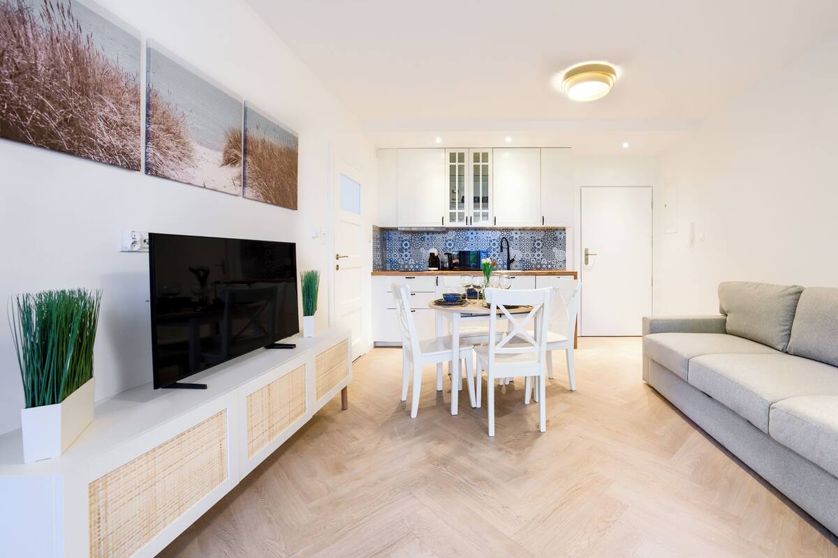 Un salon moderne avec un téléviseur à écran plat, un canapé gris et un coin repas avec des chaises blanches.