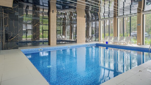 An indoor swimming pool with clear blue water, surrounded by large windows offering views of greenery and parked cars outside. - Apartamenty Sun & Snow Baltic Apartments (Pobierowo)