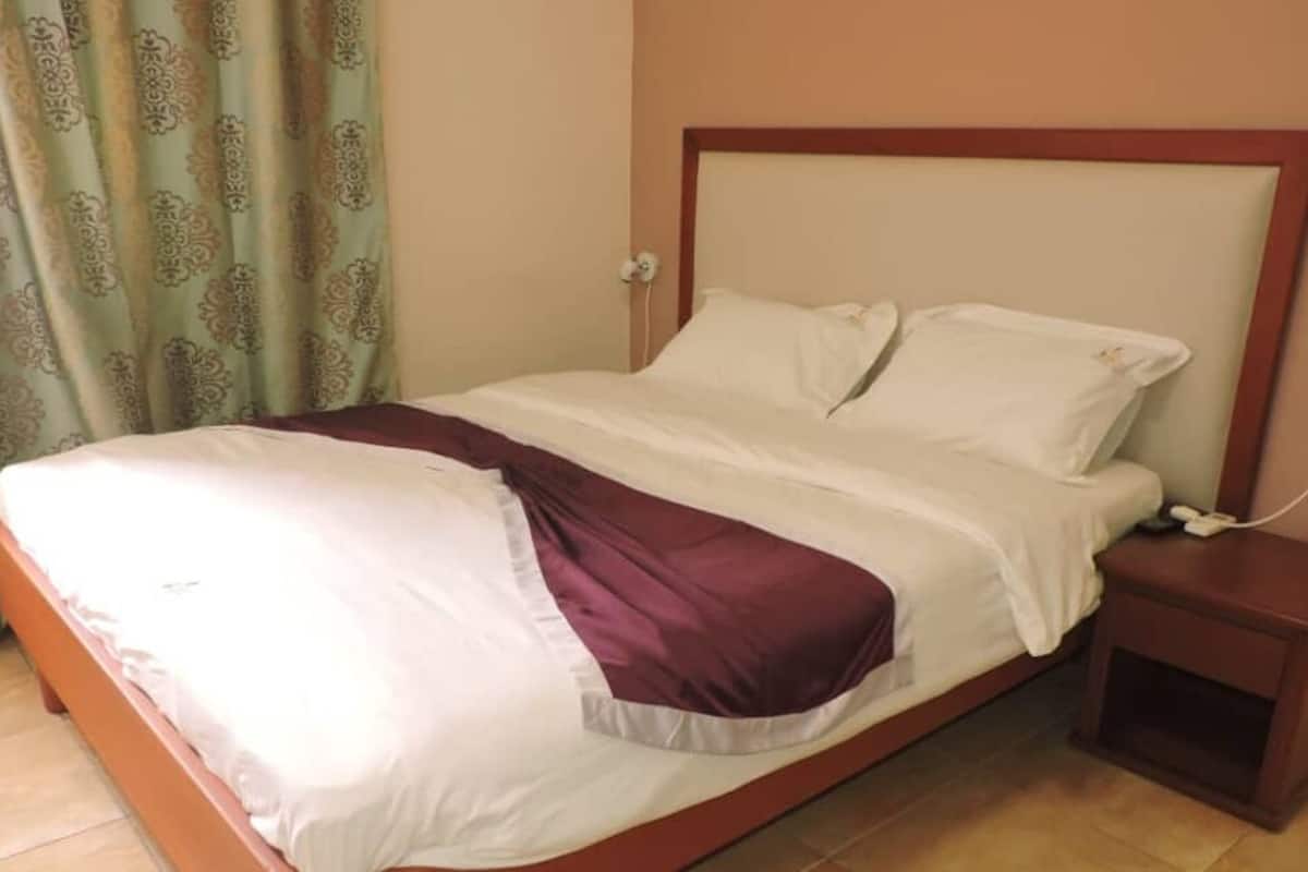 A single bed with a wooden frame and headboard, a bedside table with a phone, and a patterned curtain.
