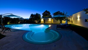 Une piscine à bord incurvé, entourée d’une terrasse dallée avec des chaises longues et des parasols. On aperçoit en arrière-plan des bâtiments éclairés de l’extérieur.
