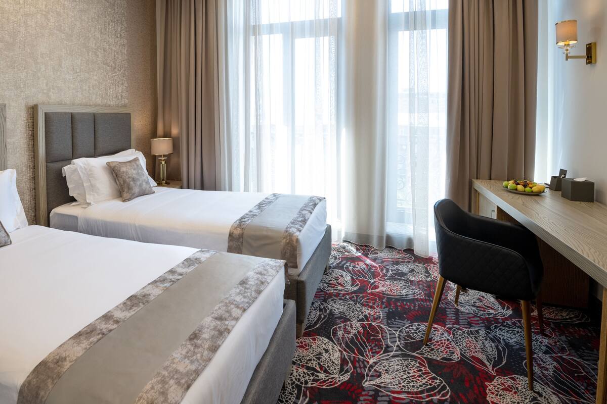 Ein Hotelzimmer mit zwei Betten, einem Schreibtisch, einem Stuhl, einer Lampe und einem Fenster mit Vorhängen.