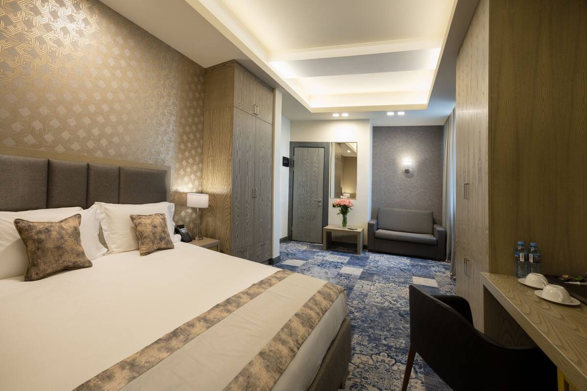 Ein Hotelzimmer mit einem großen Bett, einem Schreibtisch, einem Sessel, einem Spiegel und einem kleinen Tisch mit Blumen.