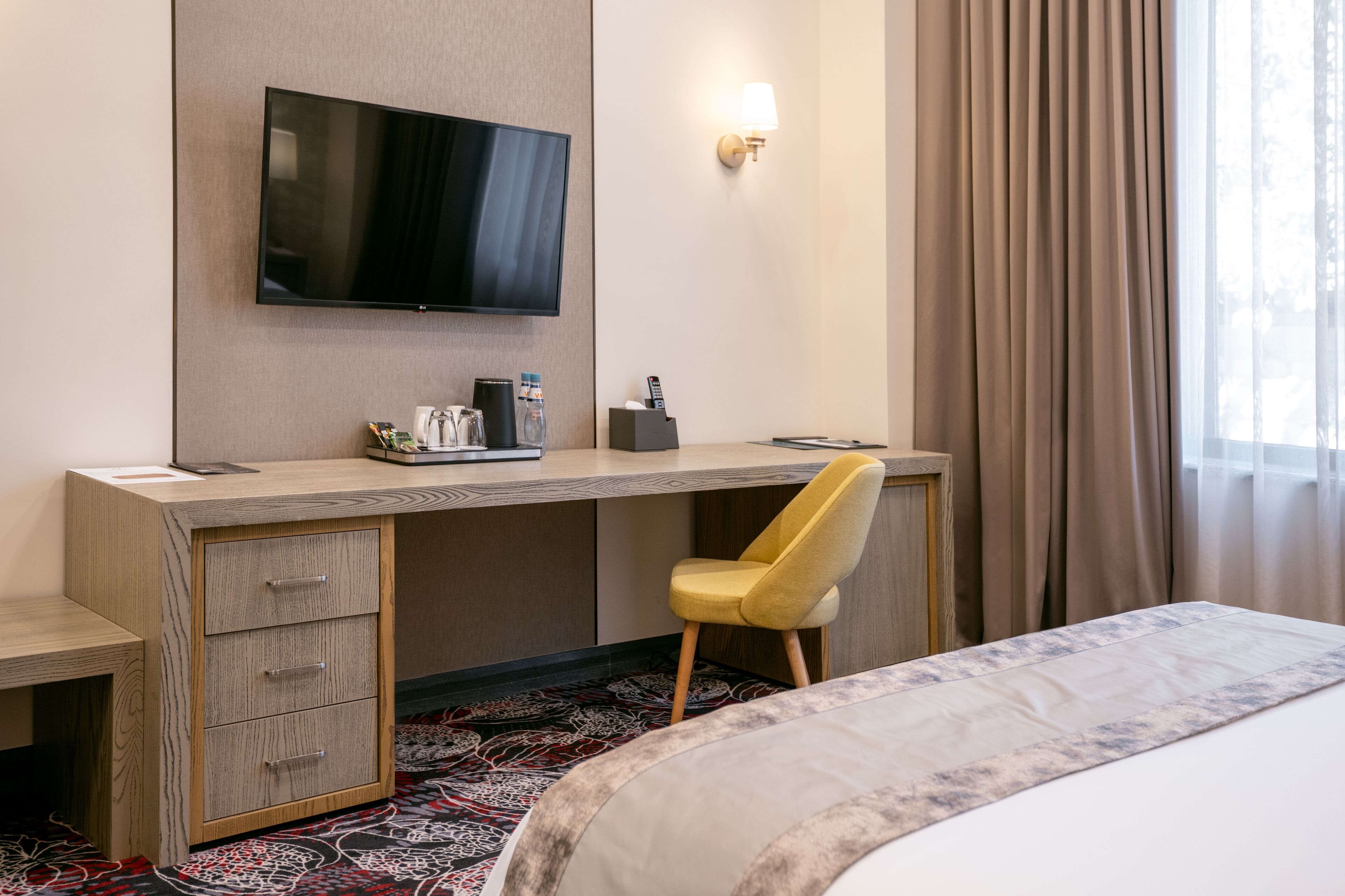 A hotel room with a wooden desk, a television mounted on the wall, a yellow chair, and a bed with a patterned bedspread.