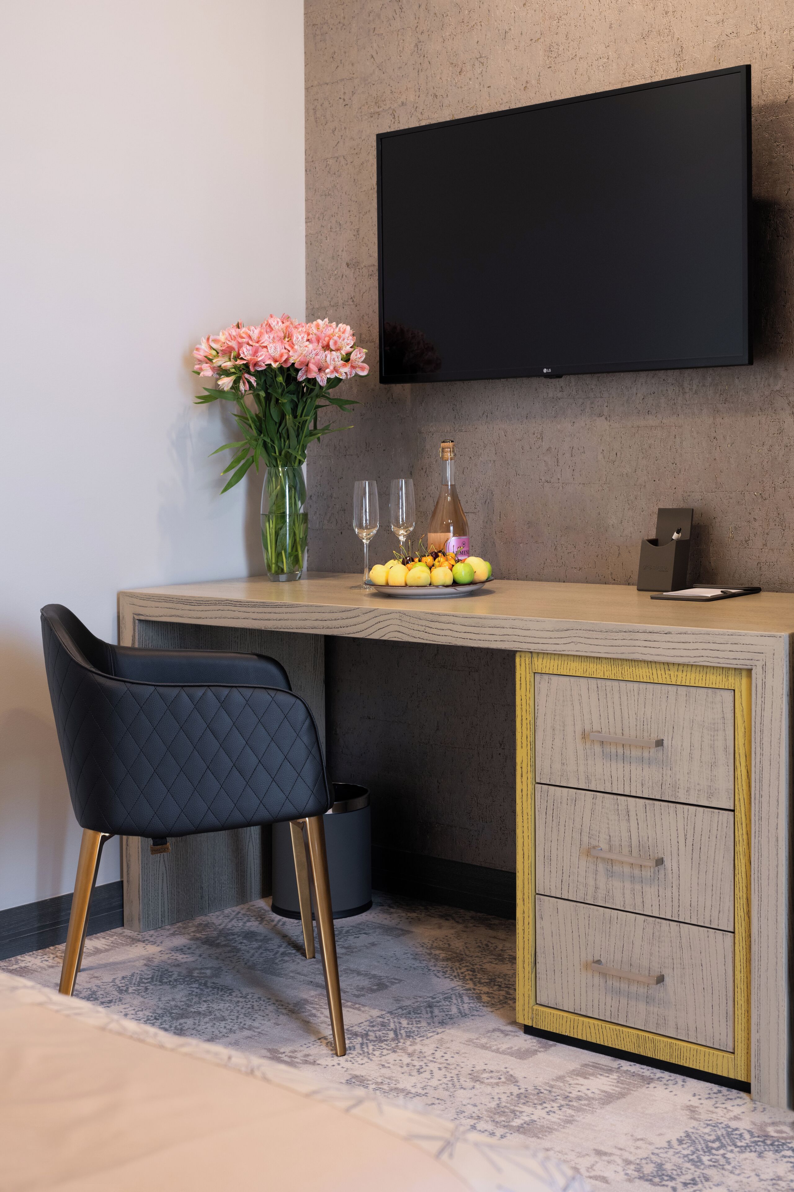 A modern room with a wooden desk, a black chair, a flat-screen TV mounted on the wall, a vase with flowers, a bottle, and a fruit bowl.