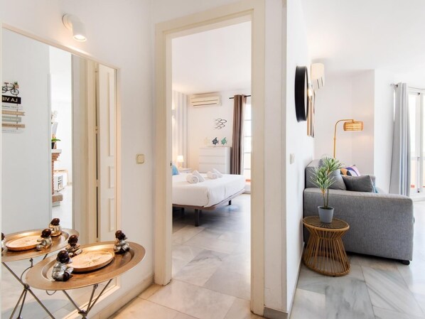 A modern hotel room with a bed, a grey sofa, a small side table with a plant, and a mirror with decorative items.