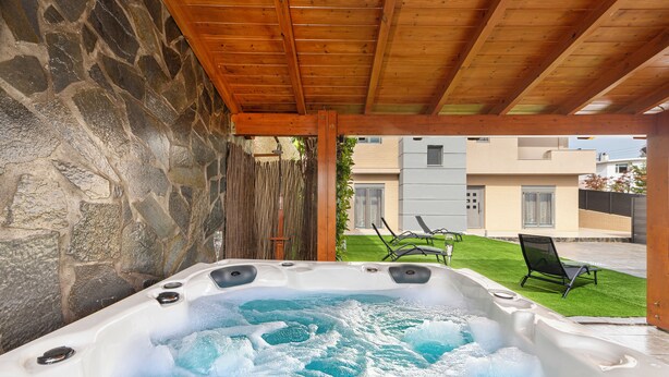 A hot tub under a wooden pergola with stone walls, a grassy lawn, and a building in the background. - ALINA HOUSE (Ialysos)