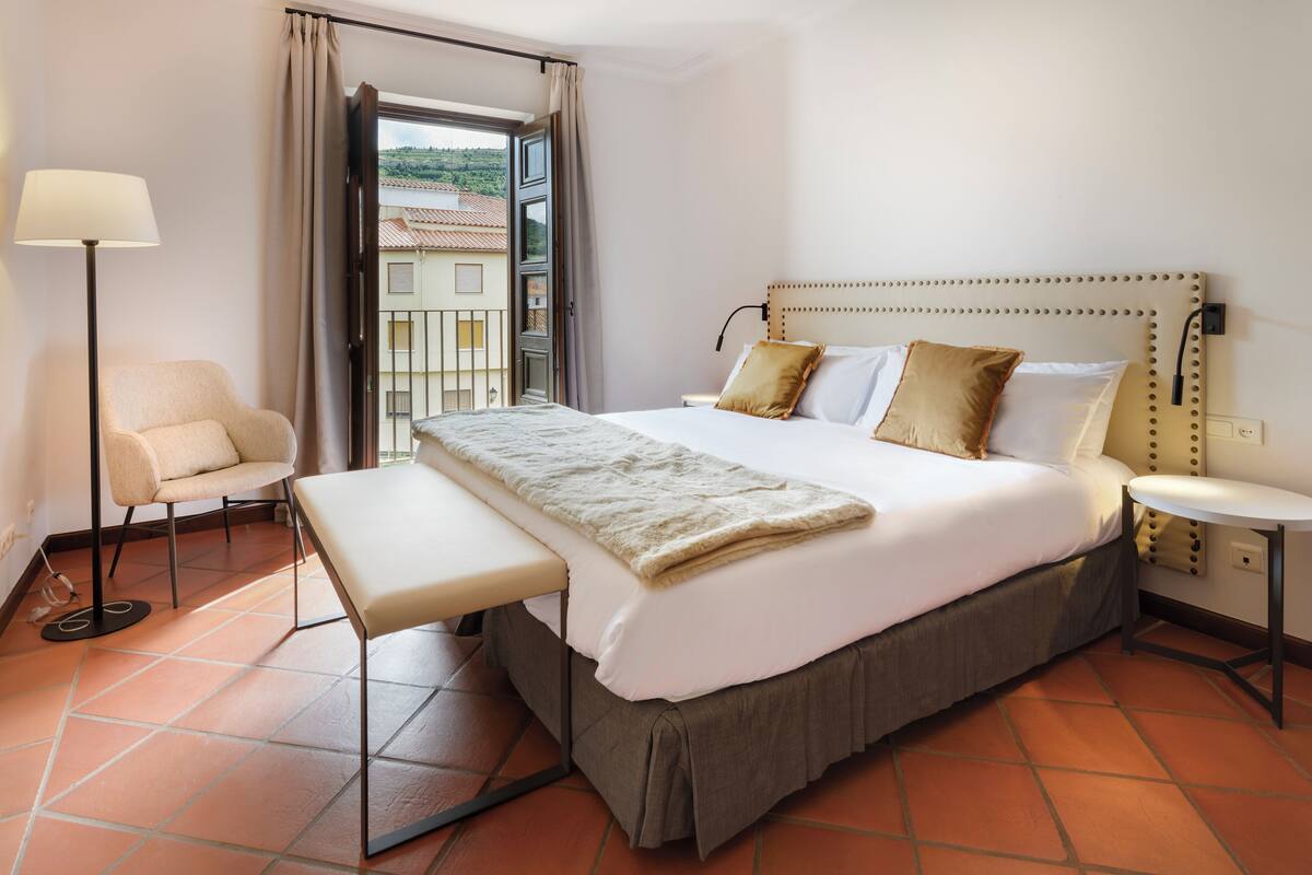 A hotel room with a bed, bedside table, lamp, chair, and an open window with a view of buildings.