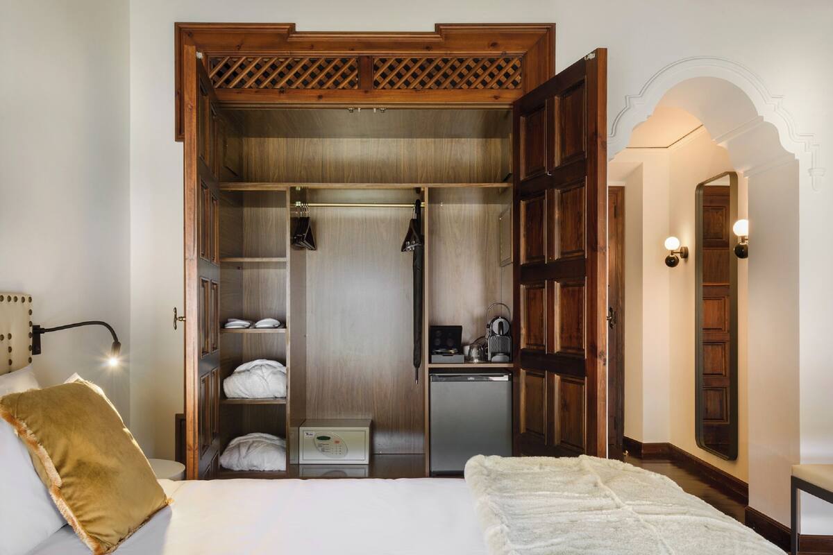 A hotel room with a bed, a wardrobe, a small kitchenette, and a mirror with wall sconces.