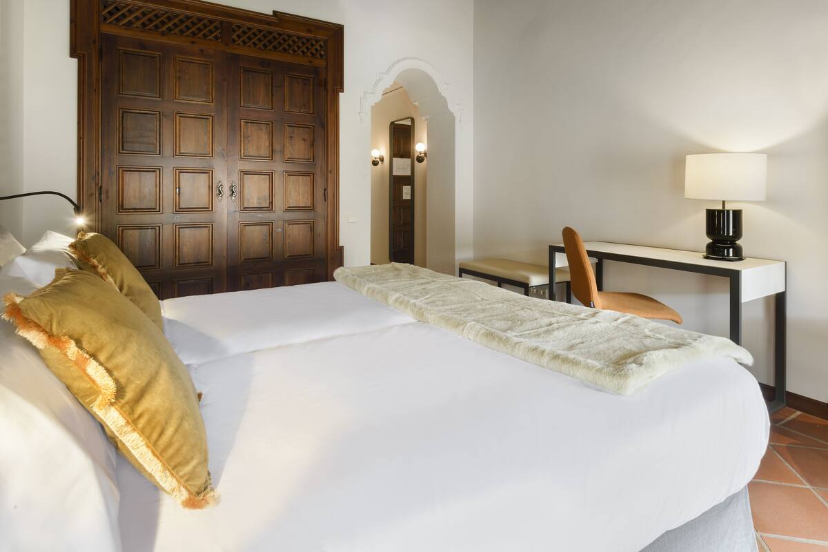 A hotel room with a large bed, a desk with a chair, a wooden wardrobe, and a mirror with wall sconces.
