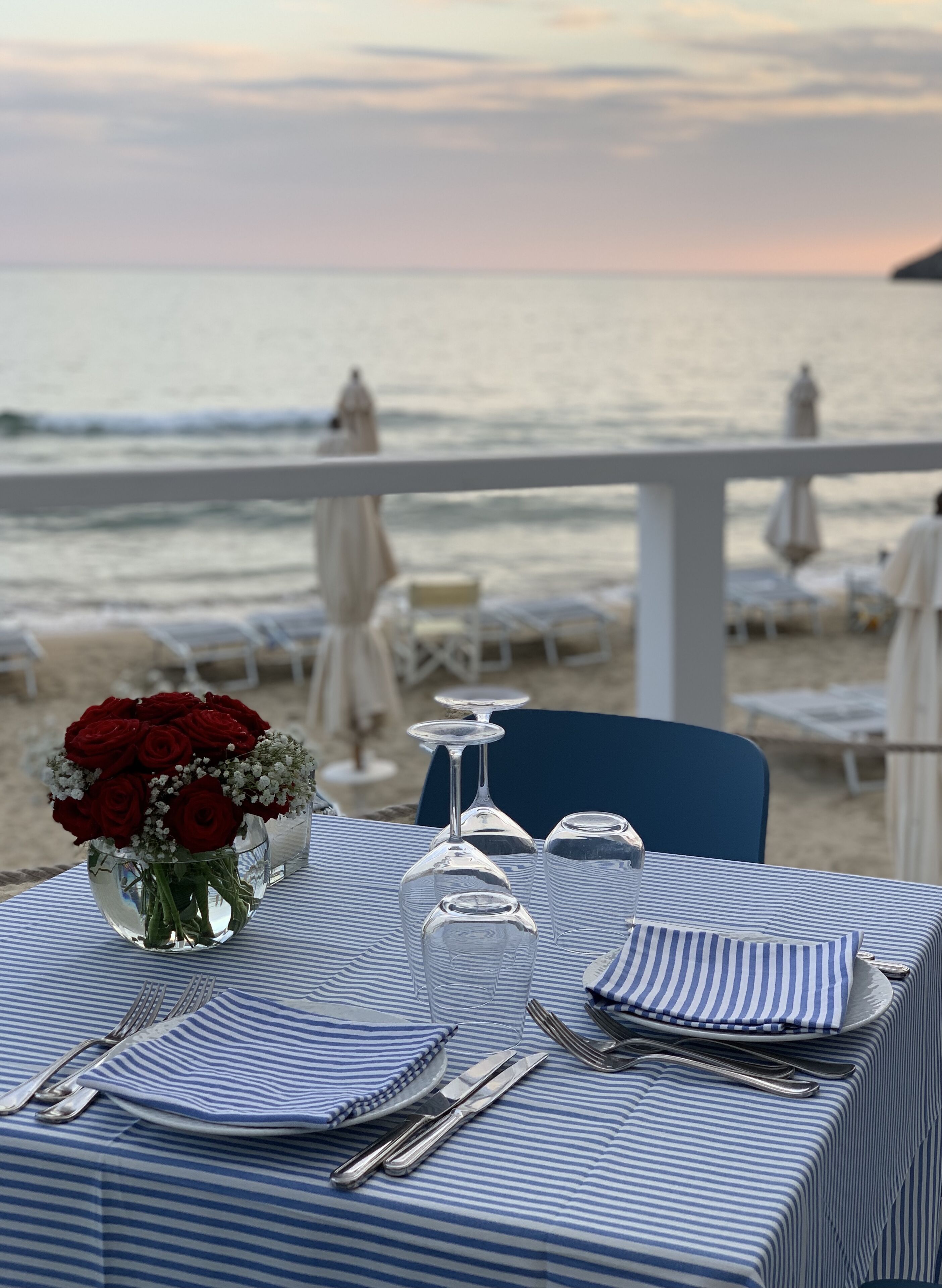 Une table à manger est dressée avec un vase de roses rouges, des verres à vin transparents et des couverts en argent, et offre une vue sur une plage parsemée de parasols et de chaises longues.