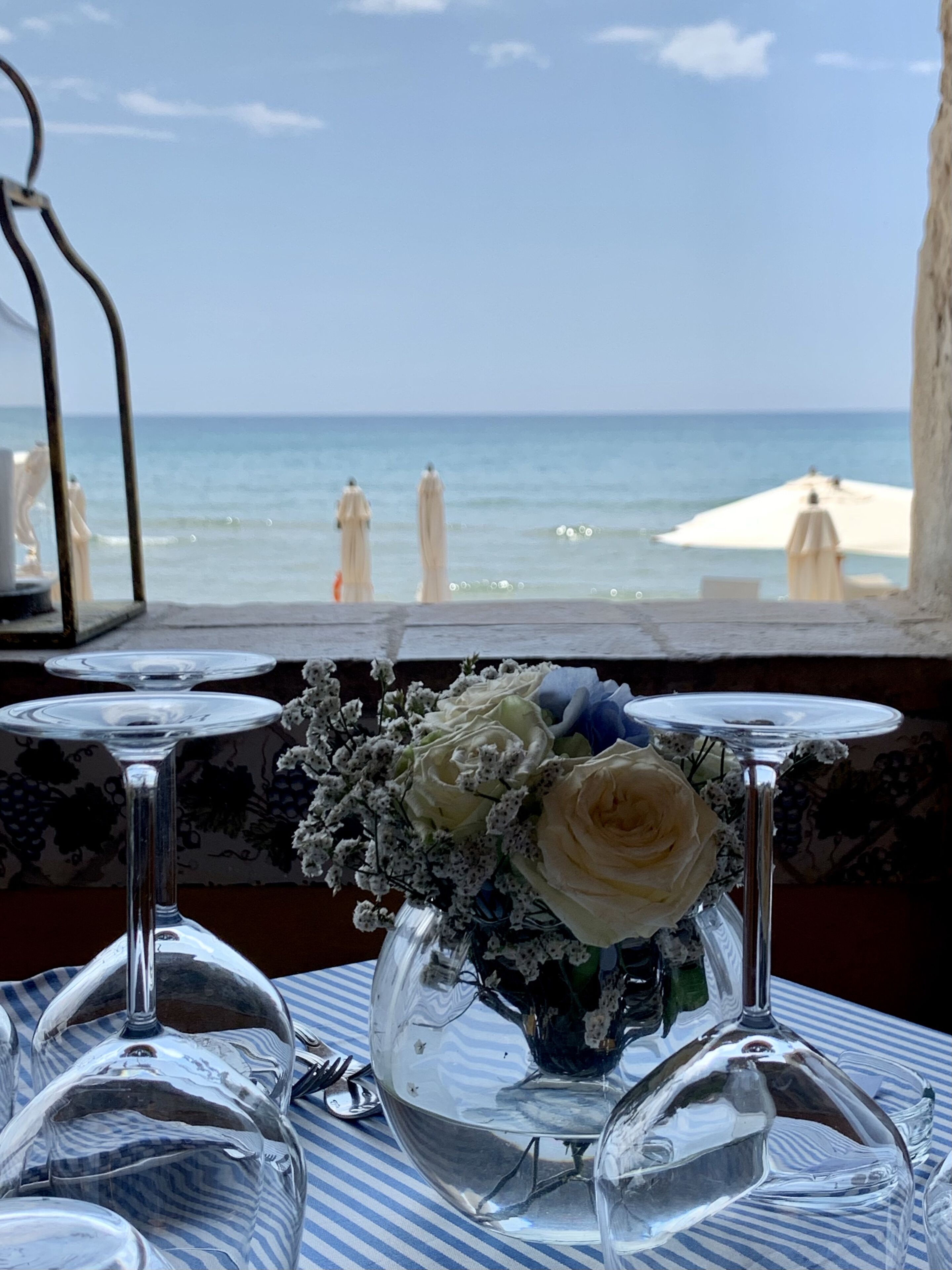 A table with empty wine glasses and a vase with flowers, overlooking a beach with umbrellas.