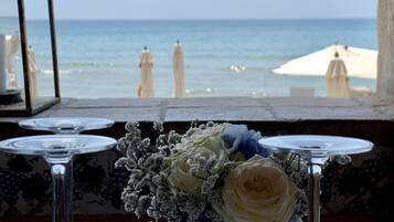 A table with empty wine glasses and a vase with flowers, overlooking a beach with umbrellas.