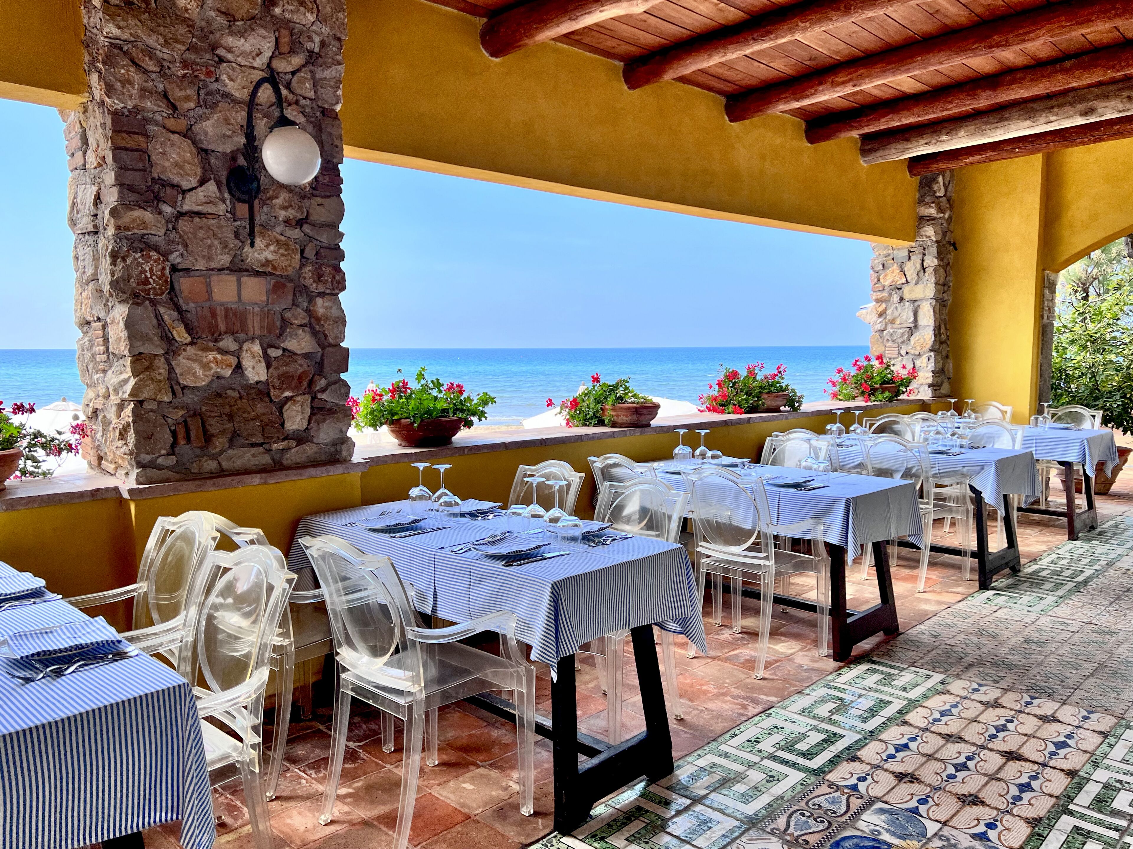 An outdoor restaurant with tables and chairs, stone pillars, and a view of the sea.