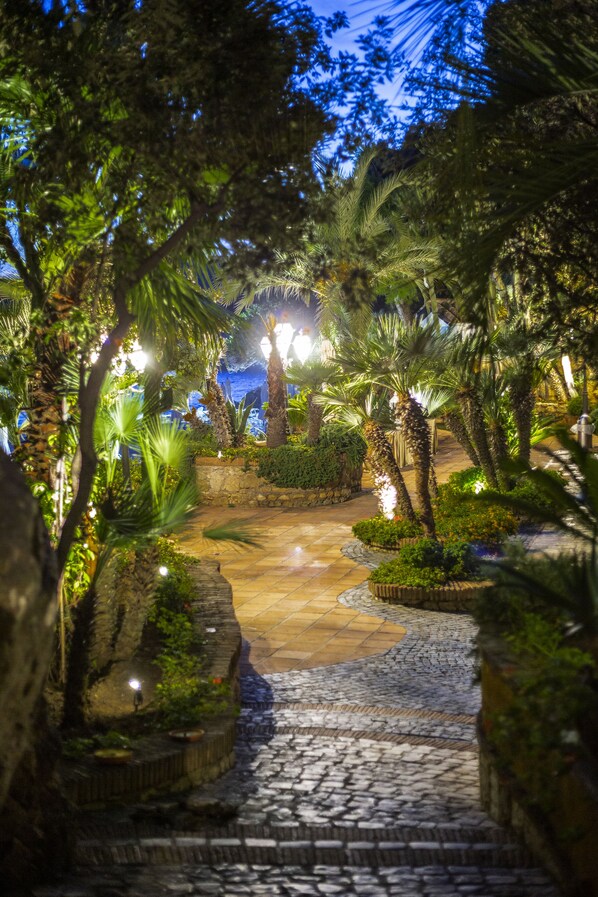 A stone pathway leading through a lush garden with palm trees and well-lit areas. - Aeneas' Landing Resort (Gaeta)