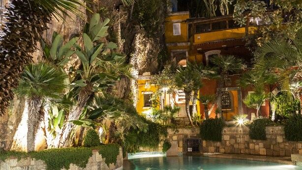 A nighttime view of a pool area with a stone wall, palm trees, and a building with illuminated windows. - Aeneas' Landing Resort (Gaeta)