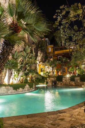 Vista nocturna de una zona de piscina con muro de piedra, palmeras y un edificio con ventanas iluminadas.