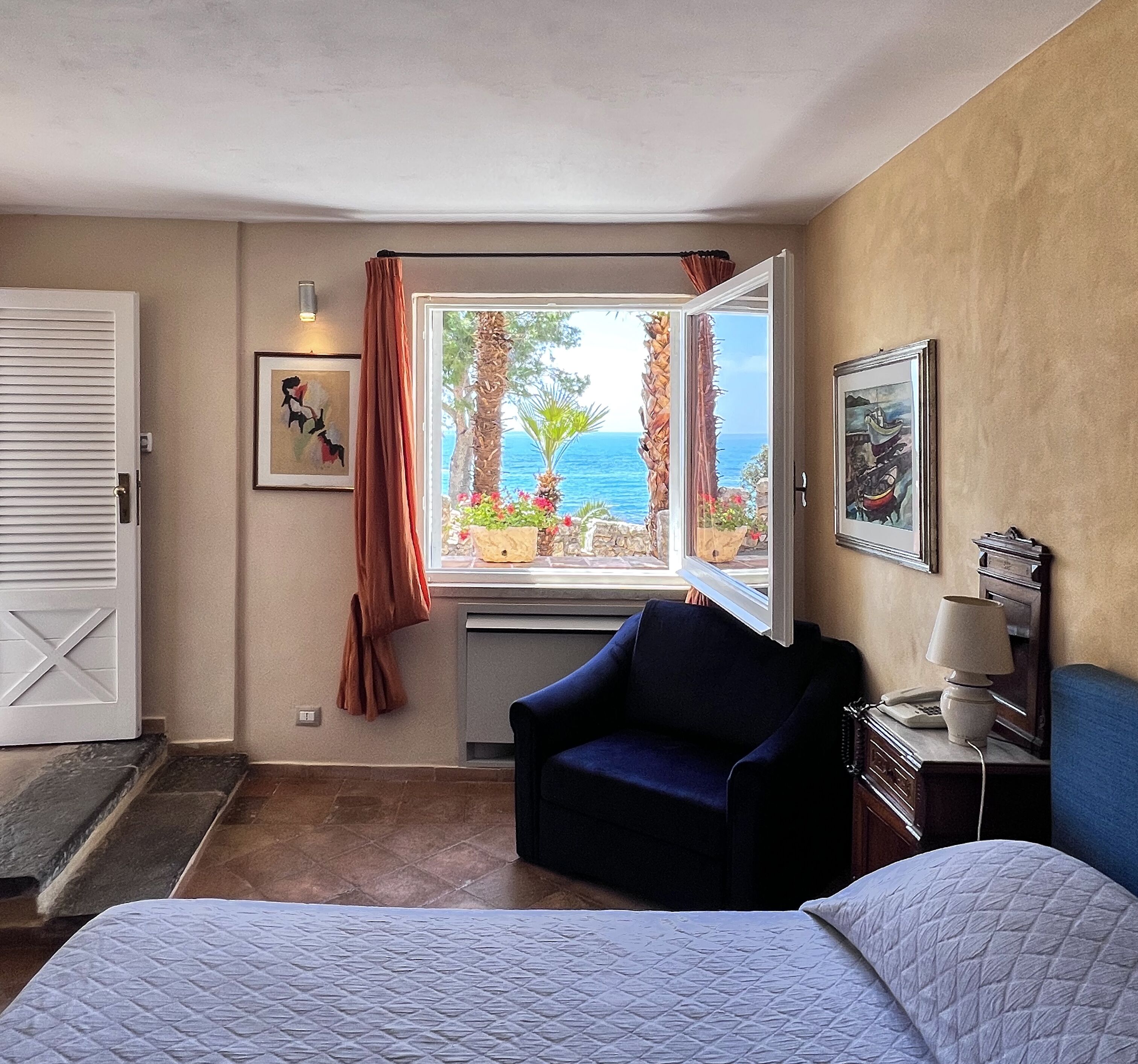 A hotel room with a bed, a chair, a desk with a lamp, a window with curtains, and artwork on the walls.