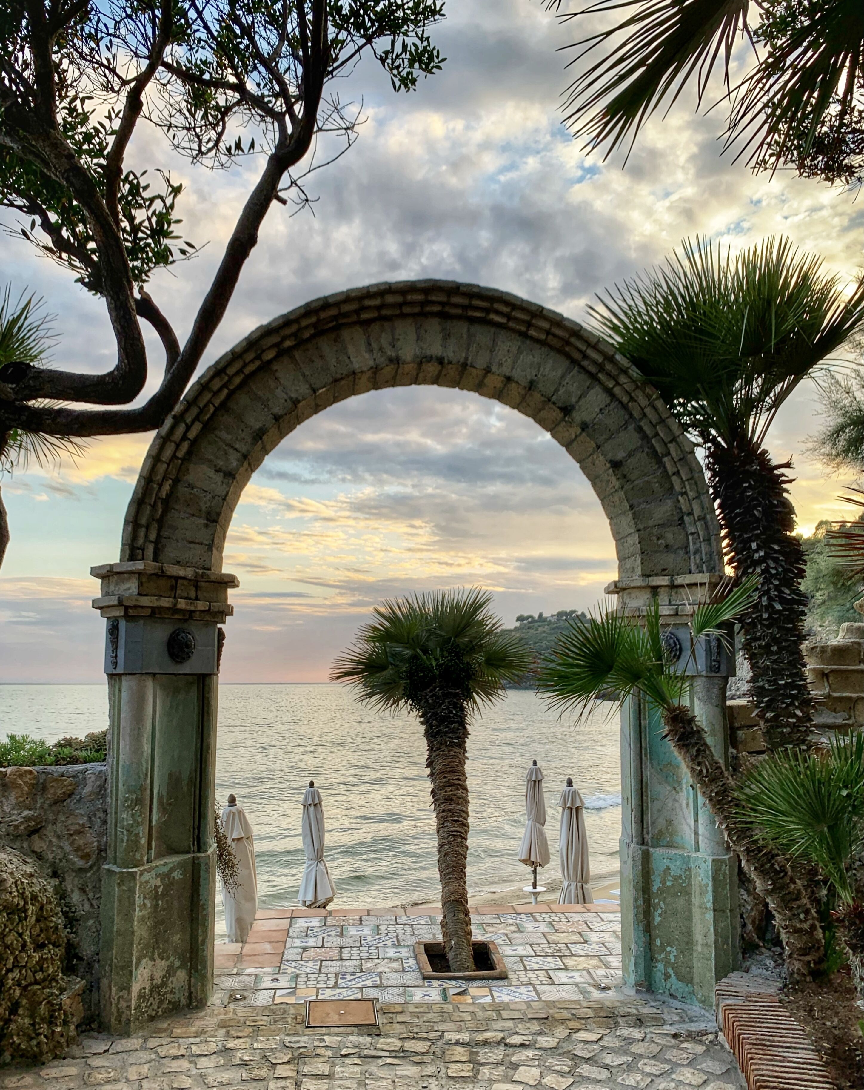 An arched stone gateway leading to a beach with umbrellas and palm trees.