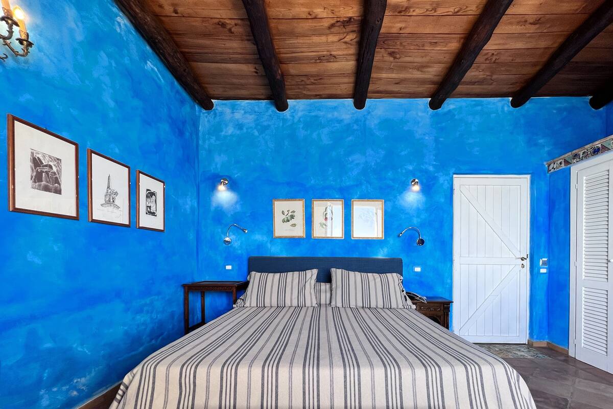 A bedroom with a blue wall, a wooden ceiling, a bed with striped bedding, and framed pictures on the wall.
