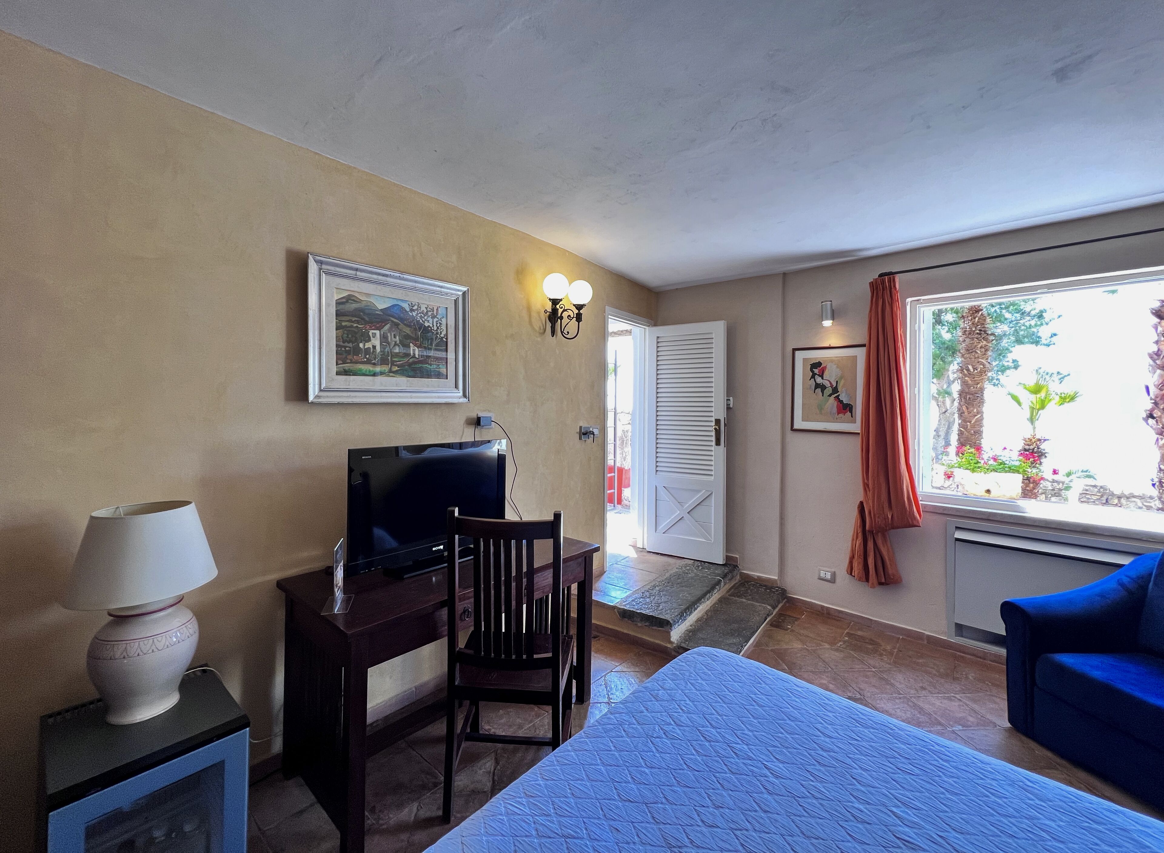 Ein Hotelzimmer mit Bett, Schreibtisch mit Fernseher, Stuhl, Lampe und Fenster mit Blick auf Grünflächen.