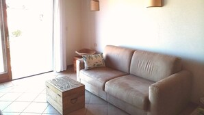 Sala de estar com sofá bege, cômoda de madeira e pequena mesa com almofada. Há duas luminárias fixadas na parede e uma porta de vidro deslizante que dá para área externa.