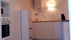 Uma cozinha compacta com armários brancos, forno embutido, geladeira e aquecedor de água fixado na parede.