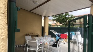 Overdekt terras met een tafel en stoelen, een droogrek en uitzicht op een zwembad en huizen op de achtergrond.