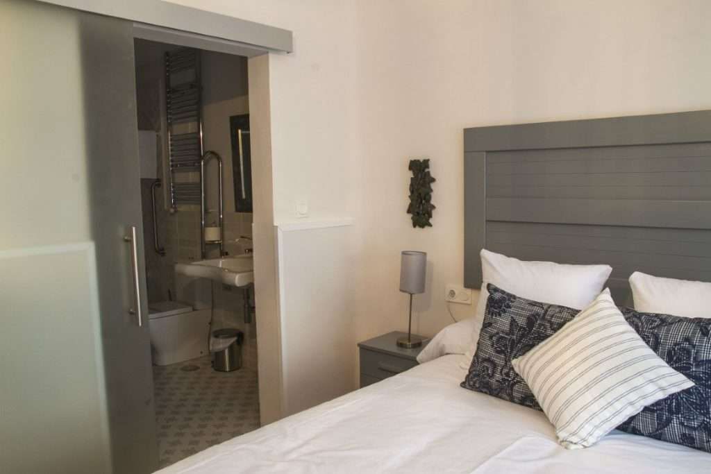 Habitación de hotel con cama, mesita de noche con lámpara, baño visible a través de una puerta abierta y una obra de arte colgada en la pared.