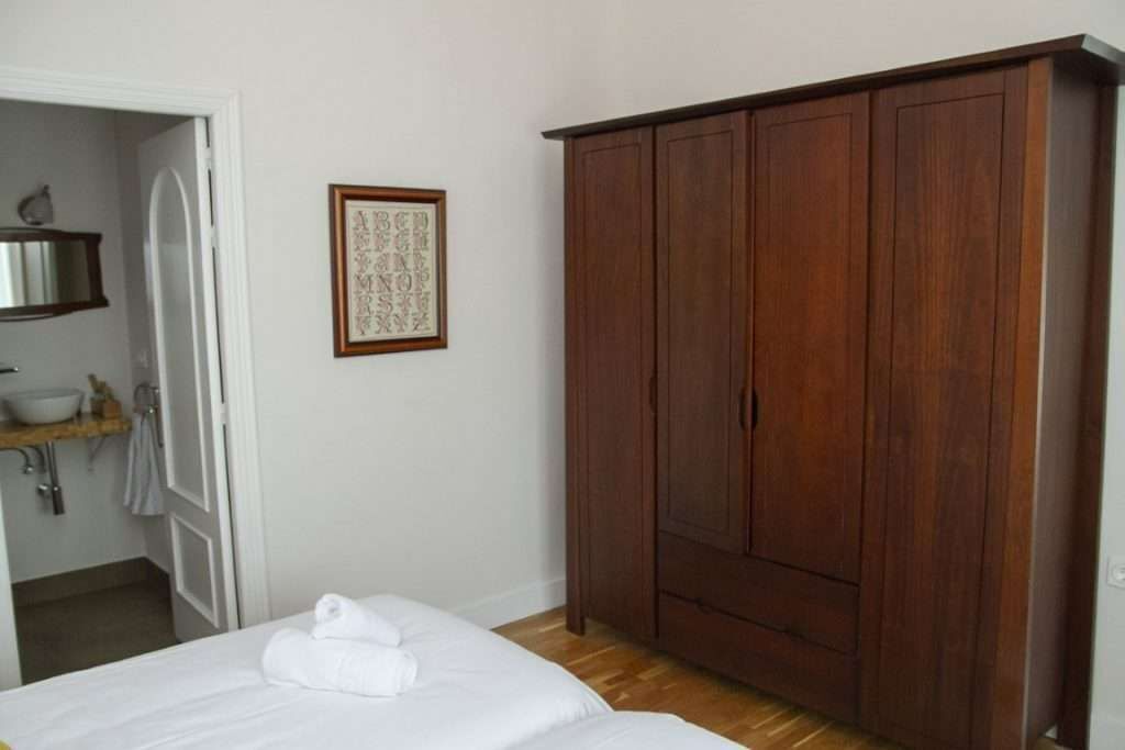 A bedroom with a wooden wardrobe, a bed with white linens, a framed picture on the wall, and a bathroom visible through an open door.