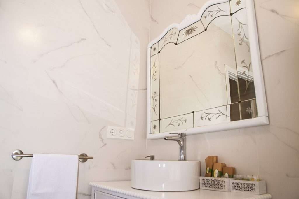 Un baño con pared de mármol blanco, un lavamanos blanco con un solo grifo, un espejo decorativo con motivos florales y un perchero con una toalla blanca.