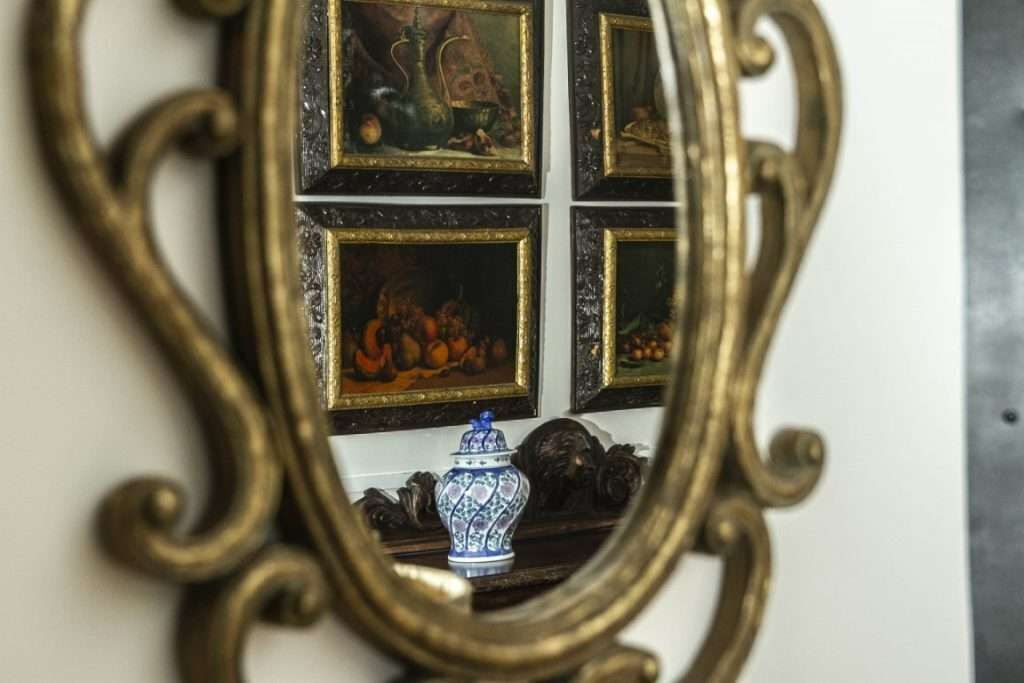 Uno specchio con cornice dorata finemente decorata riflette quattro quadri incorniciati e un oggetto in ceramica blu e bianca su una superficie scura.