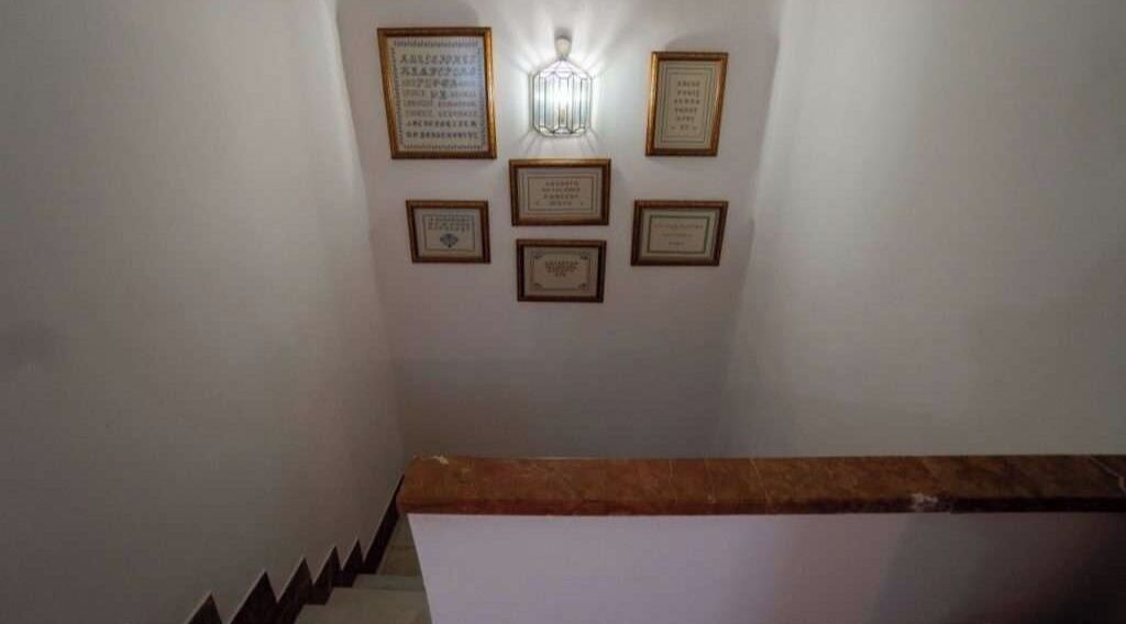 Escadaria com corrimão de madeira e parede exibindo vários certificados ou placas emolduradas.