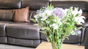 Ett vardagsrum med en mörk lädersoffa, en brun kudde och en genomskinlig glasvas med en bukett blommor på ett träbord.