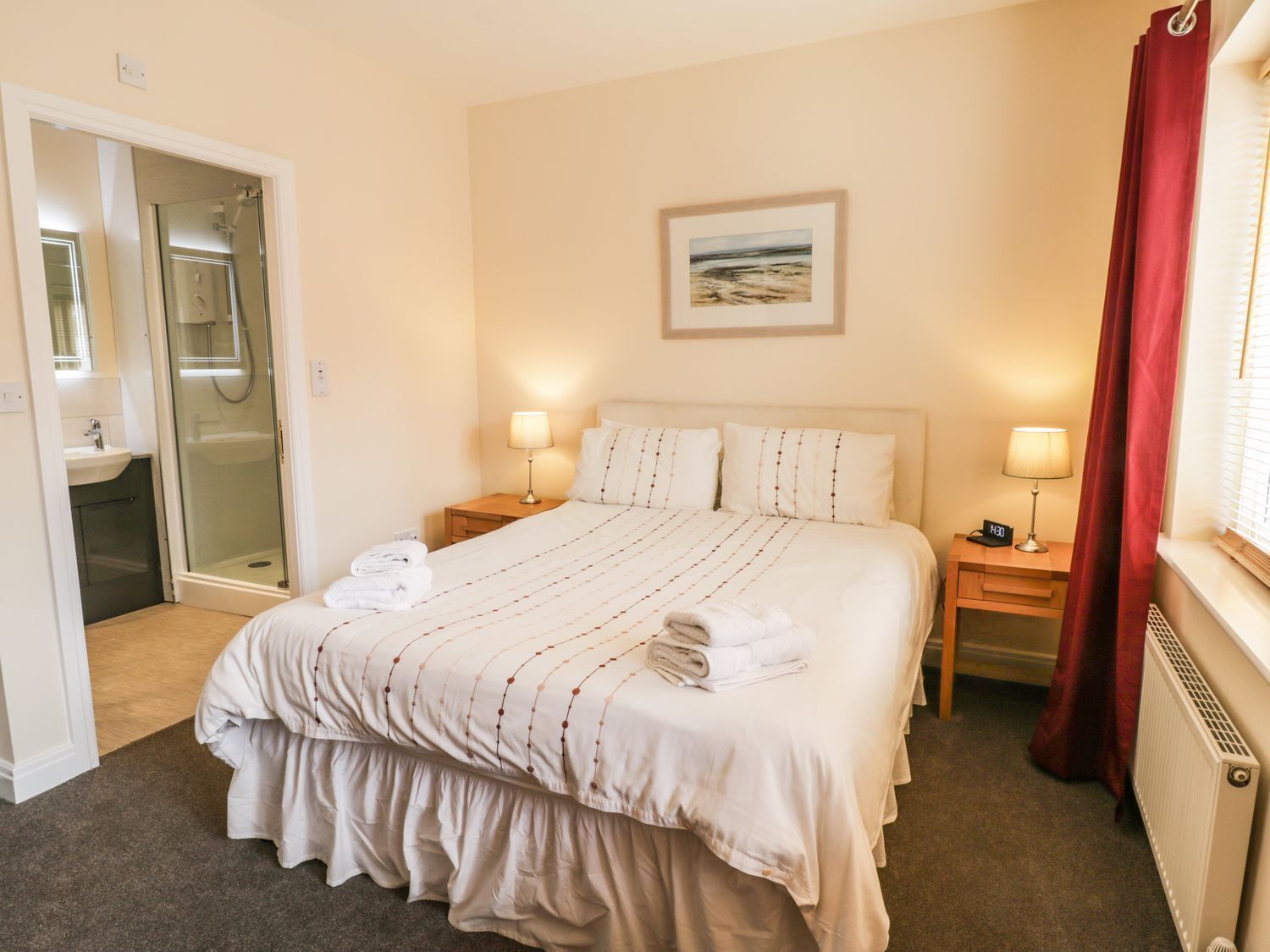 Une chambre d’hôtel comprenant un lit, deux tables de chevet, une commode, une fenêtre avec des rideaux rouges, un tableau au mur et une salle de bain avec une cabine de douche en verre.