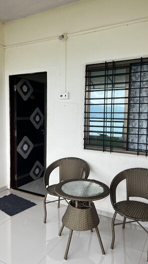 A room with a white tiled floor, a black door with a geometric pattern, a window with a metal grid, and two wicker chairs around a glass table. - Hilltop Retreat (Pune)