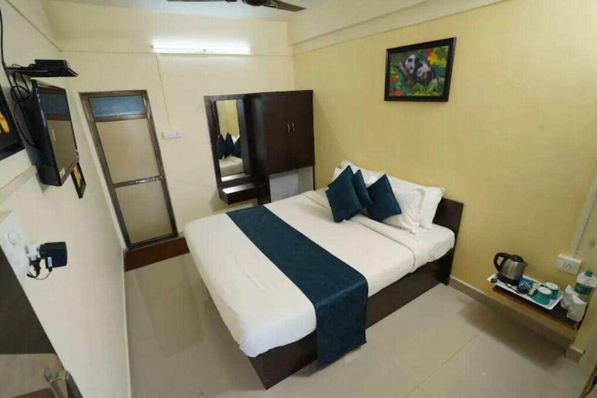 A hotel room with a bed, a wardrobe, a mirror, a painting, and a small shelf with items.