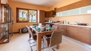 A modern kitchen with wooden cabinets, a dining table with chairs, and a view of palm trees through a window. - Harbour Views Gozitan Villa + Shared Pool (Ghajnsielem)