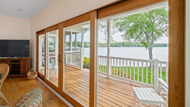 A spacious room with a large TV, wooden flooring, and a view of a lake through large sliding glass doors.