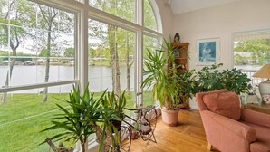 A spacious room with large windows overlooking a lake, featuring a wooden floor, a pink sofa, and various potted plants.