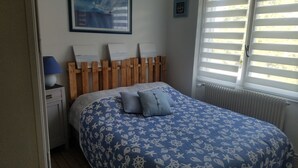 Une chambre à coucher avec une tête de lit en bois, un lit recouvert d’une couvre-lit à motifs bleus et blancs, deux oreillers, une table de chevet avec une lampe et une fenêtre avec des stores.