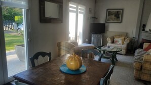 Un coin repas avec une table en bois, des chaises et un objet décoratif jaune. On aperçoit un canapé et un téléviseur à l’arrière-plan.