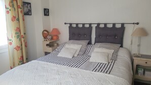 Un lit bien fait, avec une literie à rayures et deux oreillers, une table de chevet avec une lampe, et une fenêtre ornée de rideaux à motifs floraux.