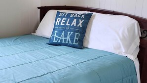 A bedroom with a bed, a blue bedspread, a pillow with text, a wall-mounted sign, and wooden flooring. - #8 2bed 1bath LAKE MI Fishing Cabin WiFi AC Deck Firepit Kayak Paddle Boat Dock (Gladstone)