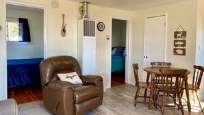 A living room with a brown leather chair, a wooden dining table with chairs, a ceiling fan, and a wall clock. - #8 2bed 1bath LAKE MI Fishing Cabin WiFi AC Deck Firepit Kayak Paddle Boat Dock (Gladstone)