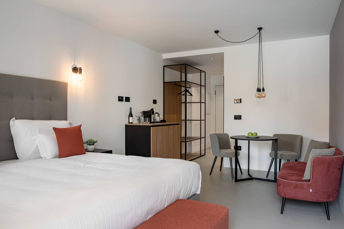 A modern hotel room with a large bed, a desk with a chair, a small table, and a hanging light fixture.